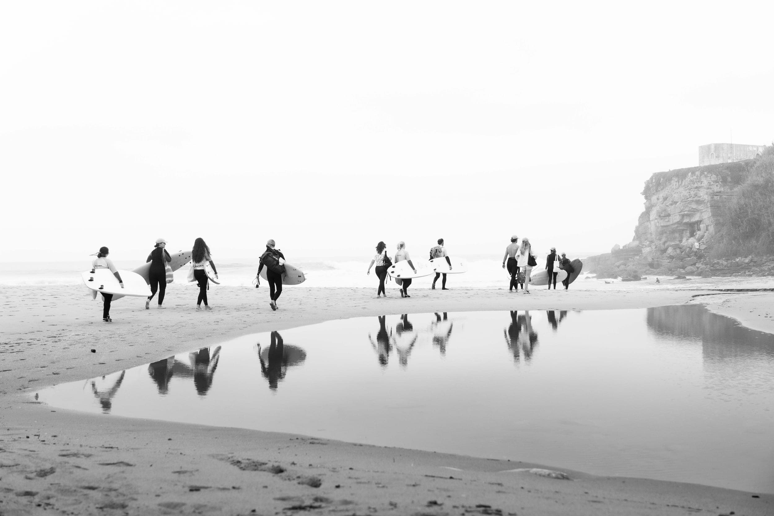 Black-and-white view of surfers carrying boards along a beach reflected in a tidal pool
