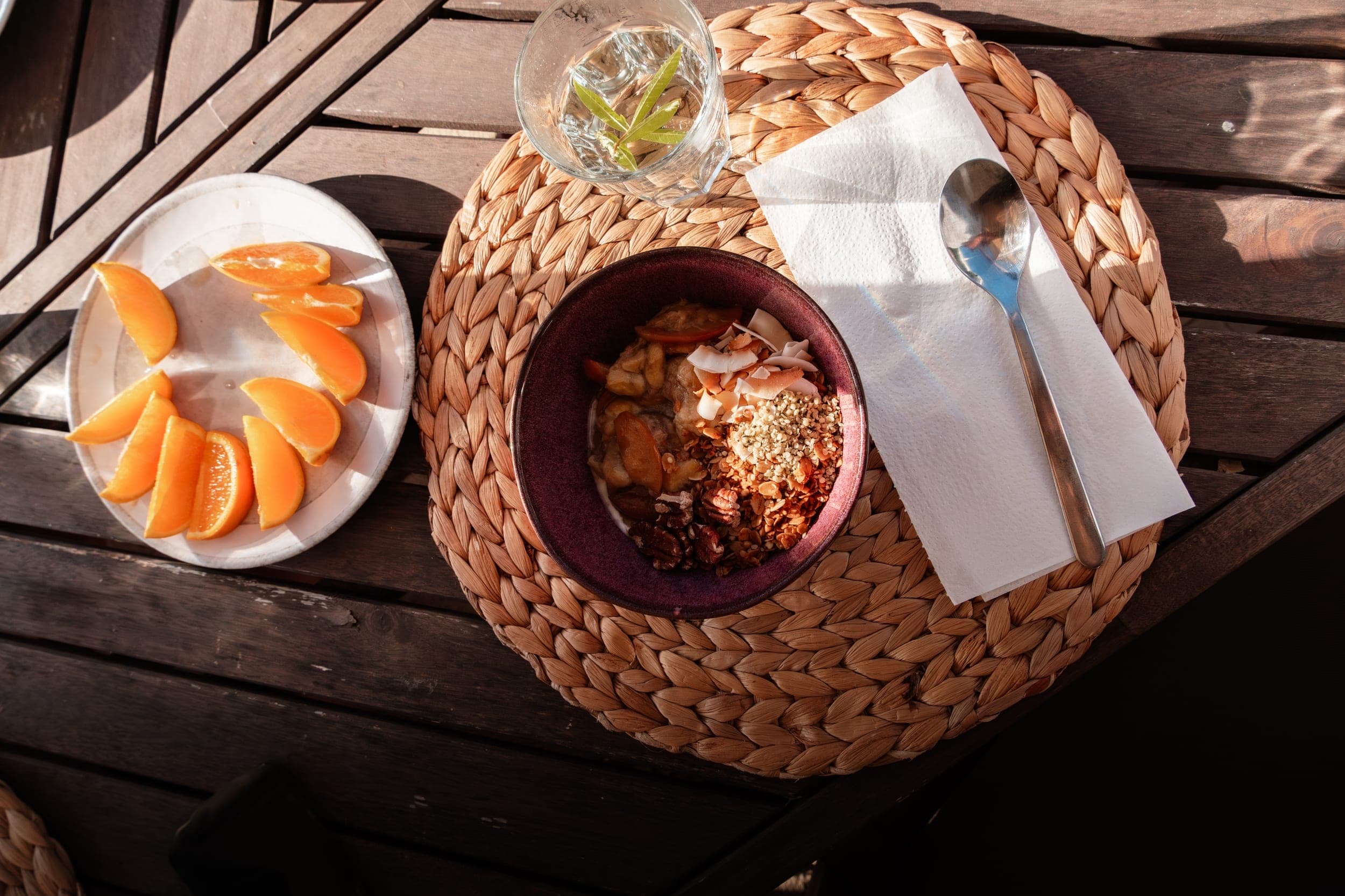Overhead breakfast of a granola bowl, orange slices and infused water on a woven placemat