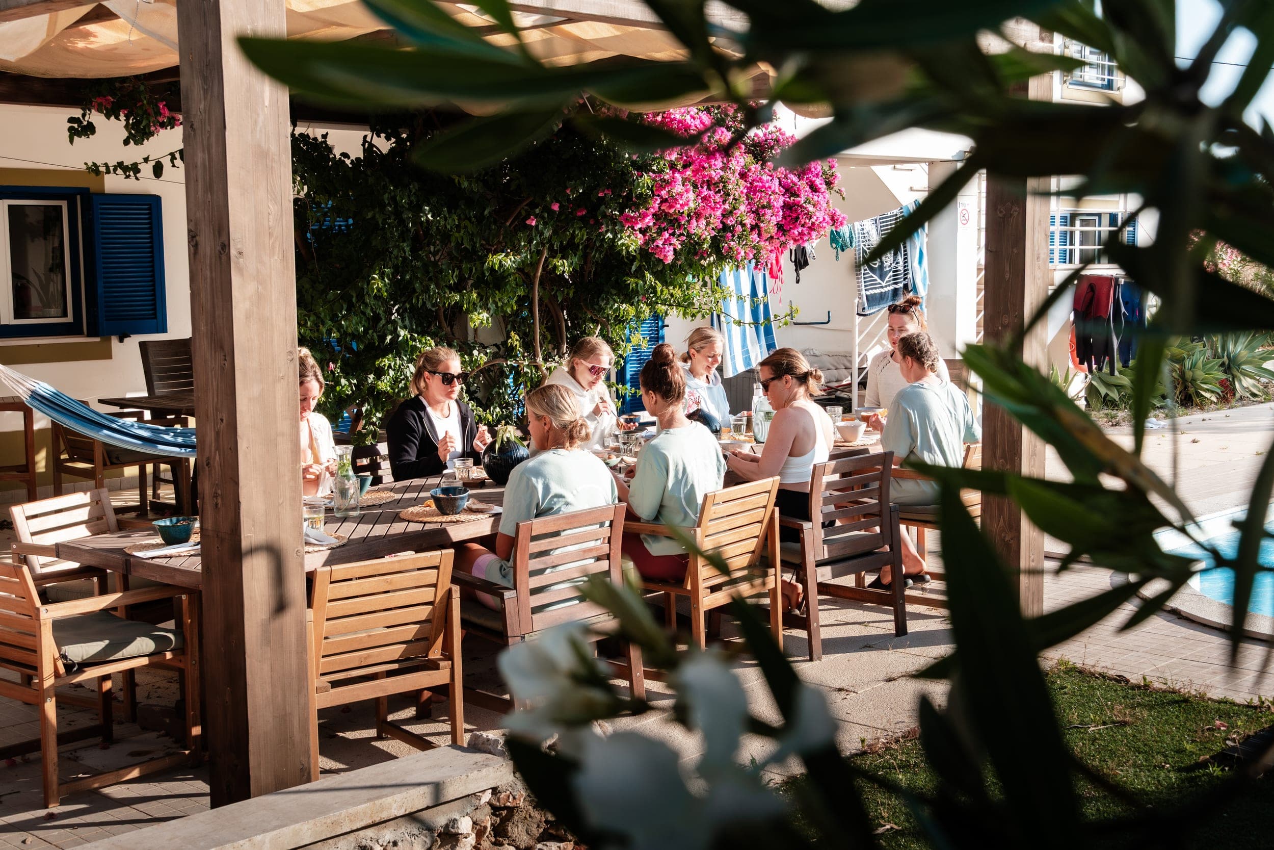 Group of women dining around a long wooden table on a shaded terrace under pink bougainvillea