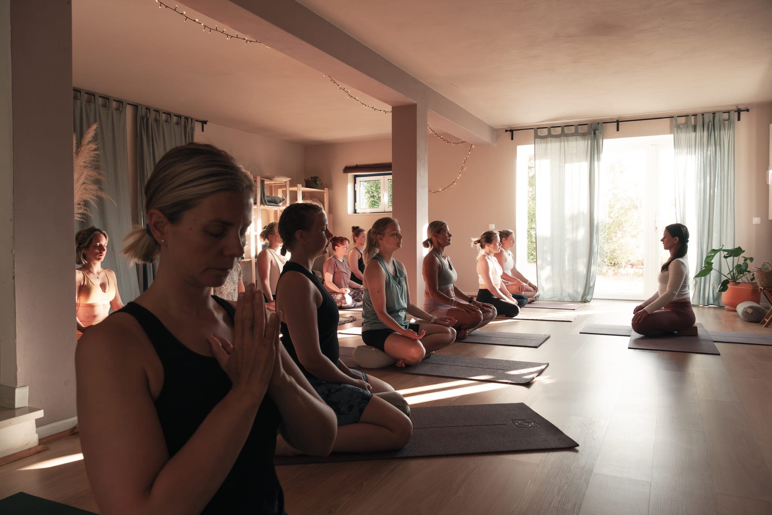 Women kneeling in meditation with hands at heart on mats in a sunlit yoga room