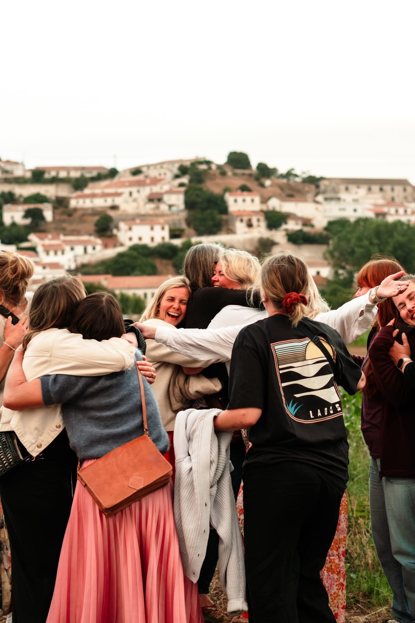 Group of women in a joyful outdoor group hug, laughing, with a hillside village behind