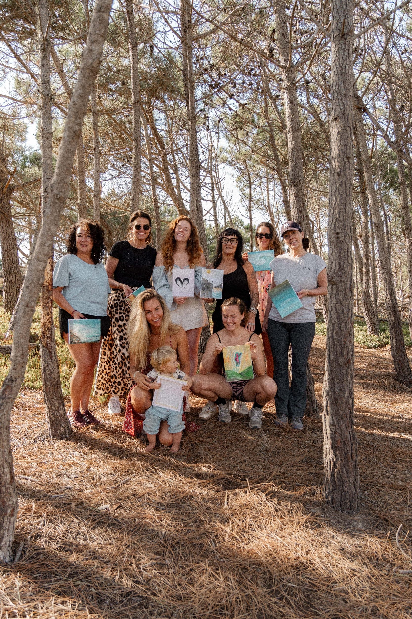 Group of women and a toddler holding painted artworks among sunlit pine trees at an Algarve retreat