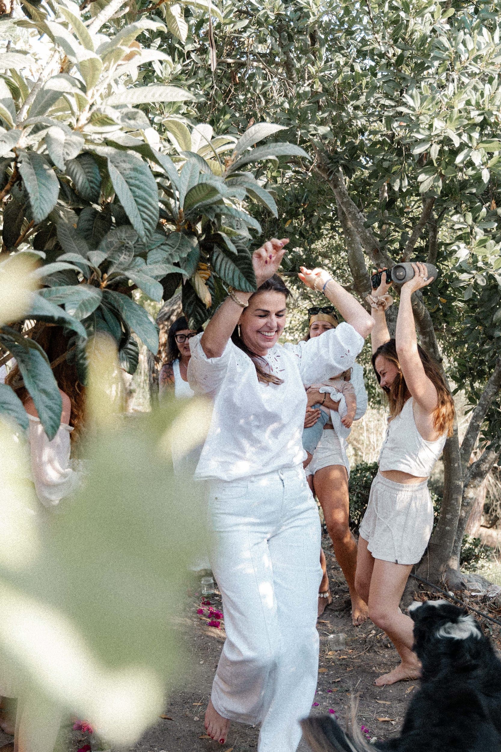 Women dressed in white dancing joyfully with raised arms under trees, a dog nearby