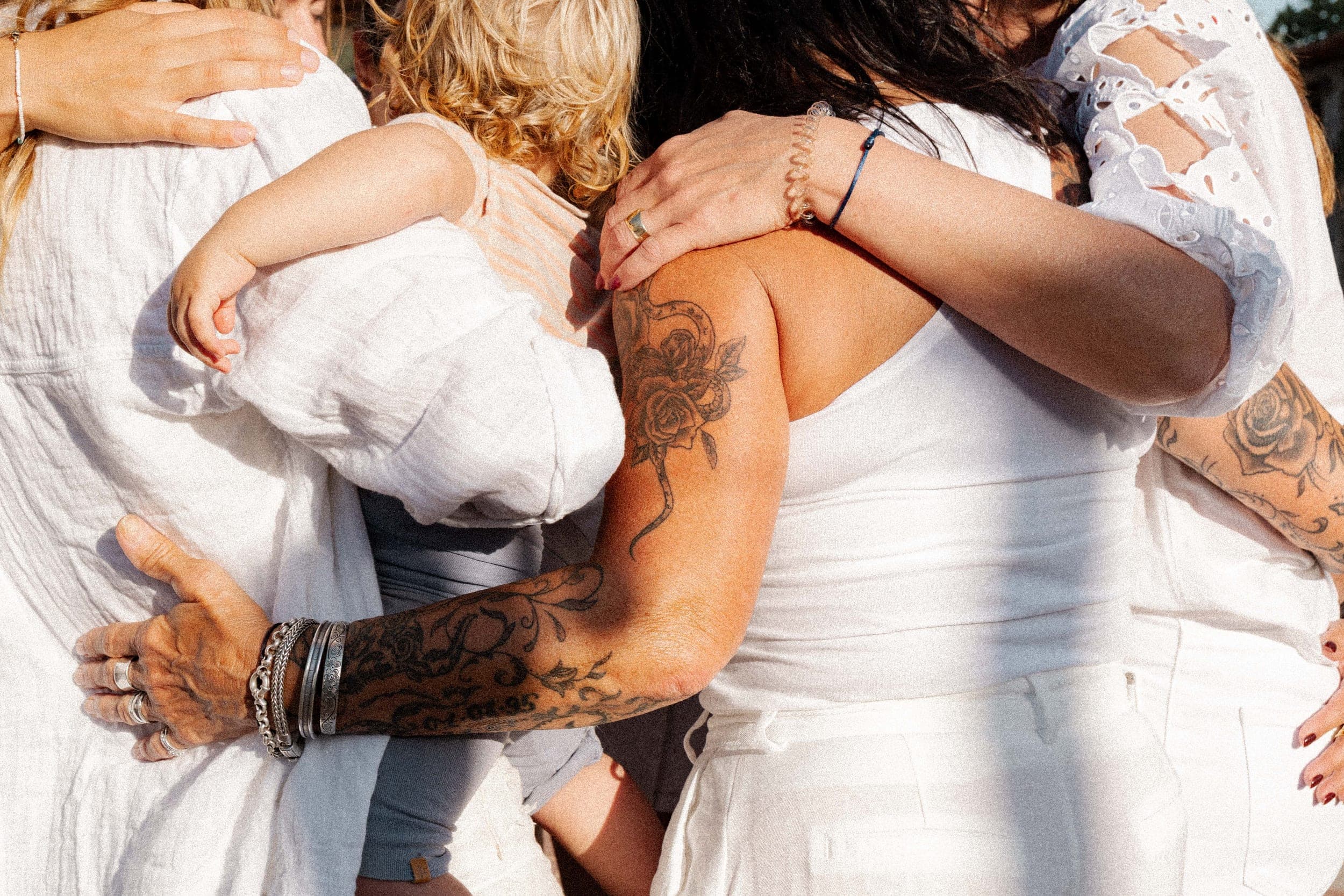 Close-up of women in white embracing in a group hug, tattooed arms wrapped around each other