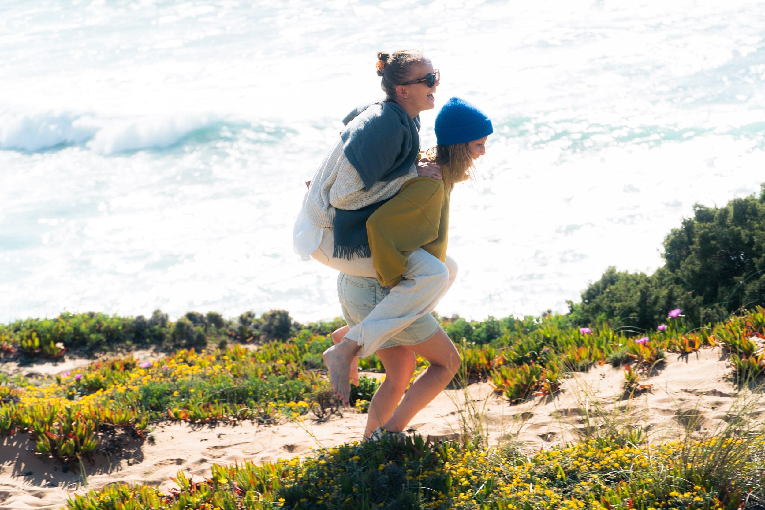 Laughing woman giving another a piggyback ride across flowering coastal dunes