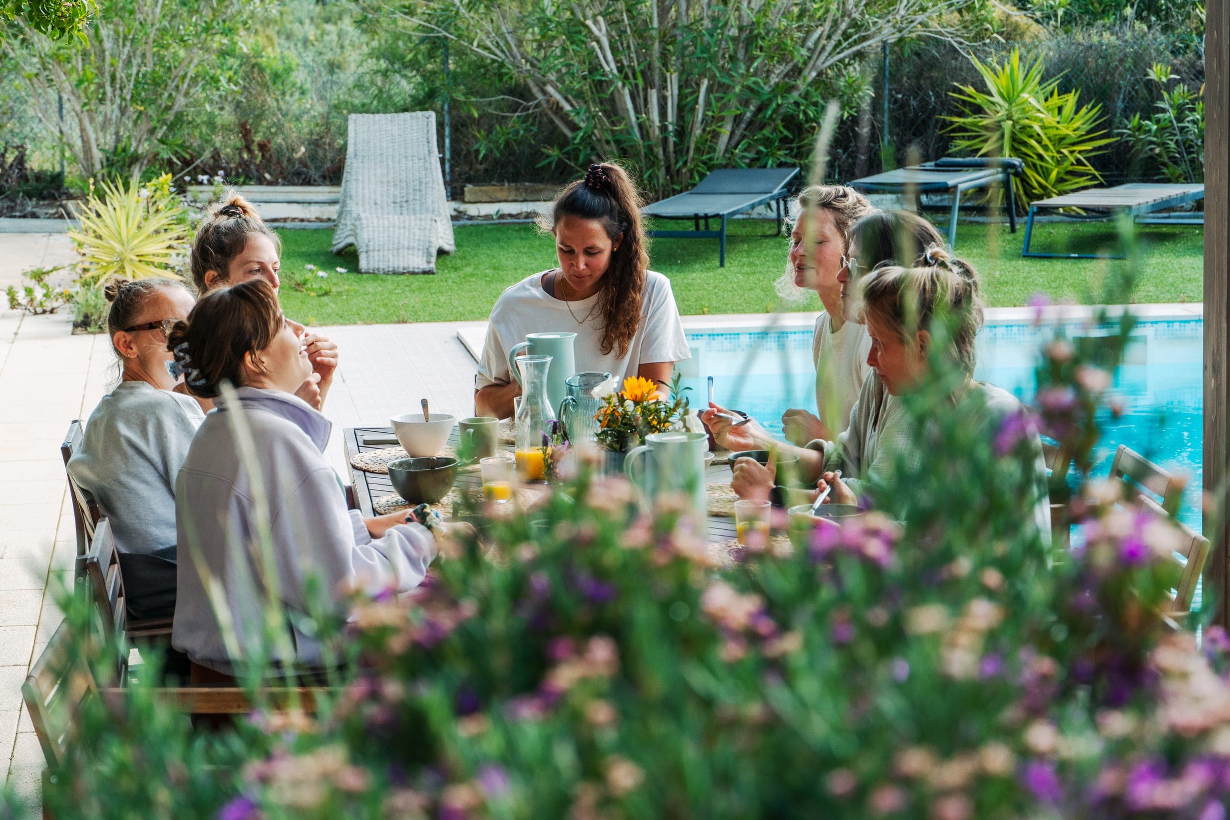 Women sharing breakfast at a poolside table framed by garden flowers