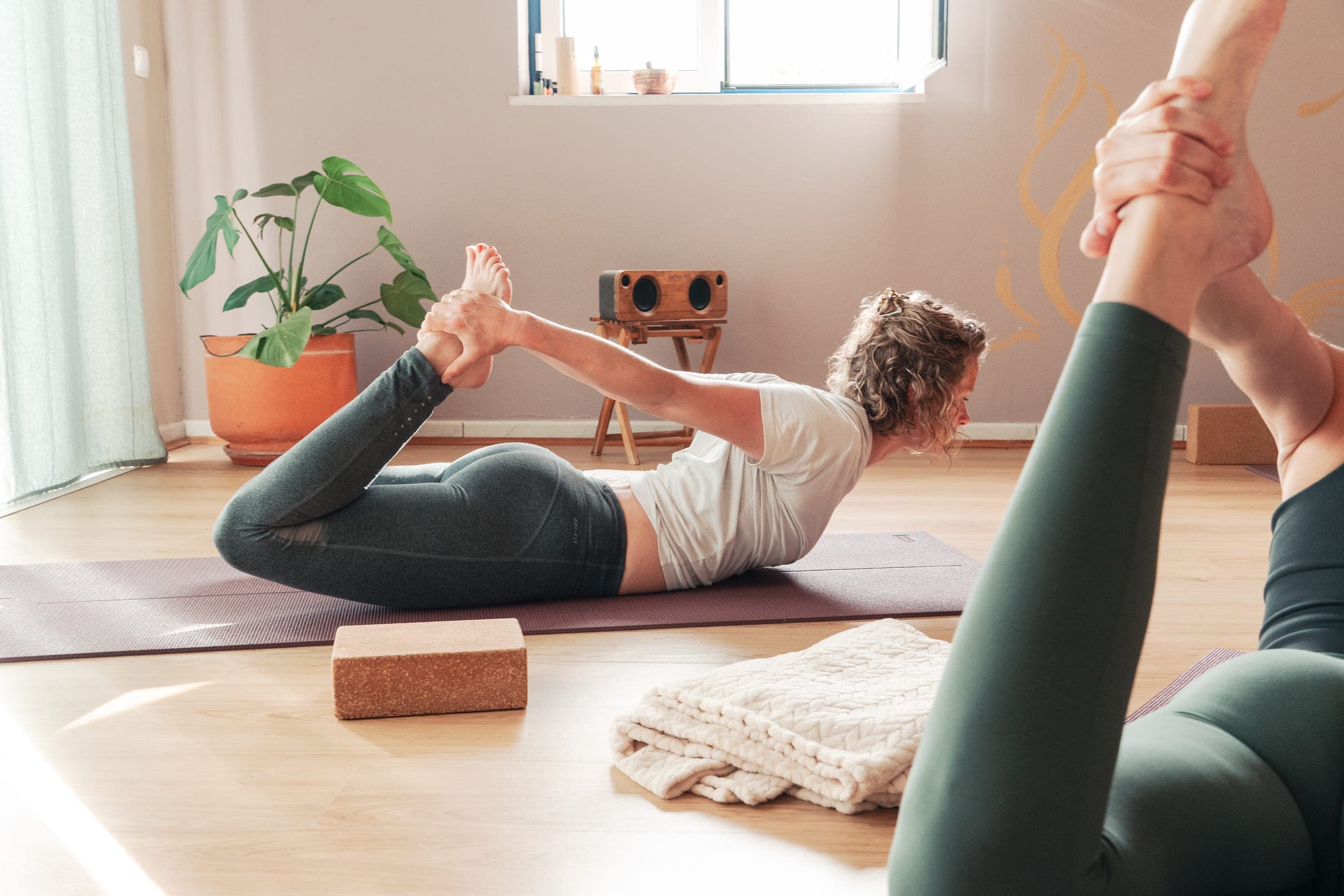 Woman in bow pose gripping her feet on a mat in a sunlit plant-filled yoga room